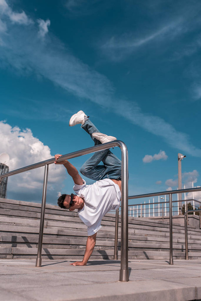 young guy a dancer in a white T-shirt, jeans, dancing break dance in summer in the city, in sunglasses, white sneakers background building clouds, active hip hop, youth lifestyle - Valokuva, kuva