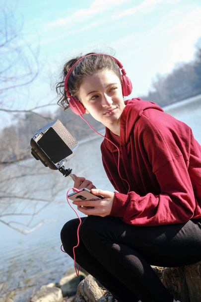  mosolygós tinédzser lány rózsaszín headphoens zenét hallgatni a mobil kívül egy tó mellett - Fotó, kép