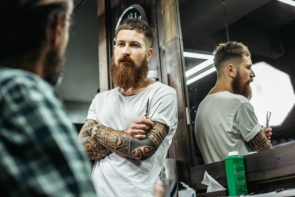 Calmo curioso barbeiro barbudo ter seus braços tatuados cruzados e olhando atentamente para seu cliente
 - Foto, Imagem