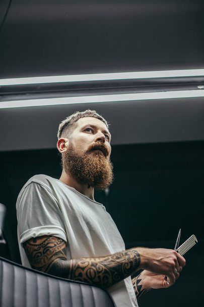Calm attractive bearded barber with tattoos on his arms thoughtfully looking into the distance while holding comb and scissors - Φωτογραφία, εικόνα
