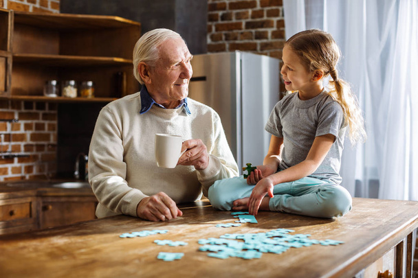 Pleasant weekend time. Cheerful elerly man assembling jigsaw puzzles with his granddaughter while resting at home - Photo, image