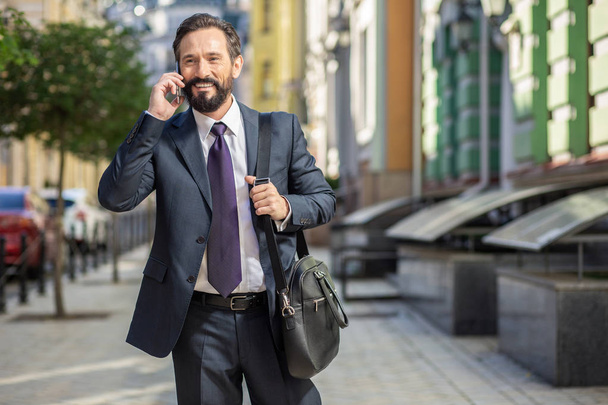 Rapid lifestyle. Cheerful handsome businessman talking on phone while walking to work - Photo, Image