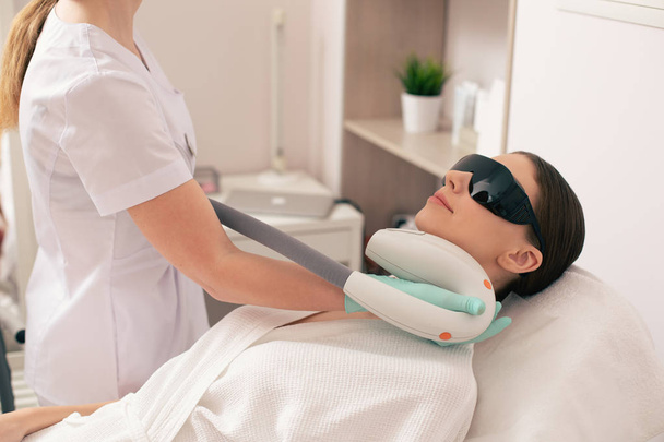 Mujer joven acostada en el sofá médico con gafas protectoras negras en la cara y sonriendo mientras se realiza un procedimiento de depilación láser
 - Foto, imagen