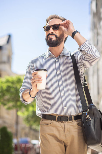 Sunny day. Delighted bearded man drinking coffee while walking along the street - Foto, imagen