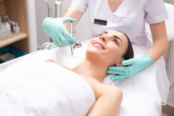 Cheerful relaxed woman lying on the medical couch and smiling while dermatologist holding a modern tool for oxygen spray treatment - Фото, изображение