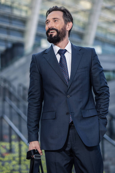 Корпоративный мир. Веселый бородатый улыбающийся бизнесмен стоит на улице, держа свою сумку
 - Фото, изображение
