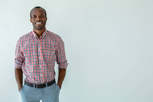 Upbeat mood. Positive smiling afro american man holding hands in the pockets while smiling against white background - Photo, Image