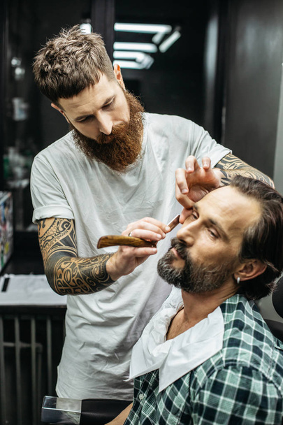 Careful professional barber with long beard standing close to his client and holding straight razor while shaving him - Fotó, kép