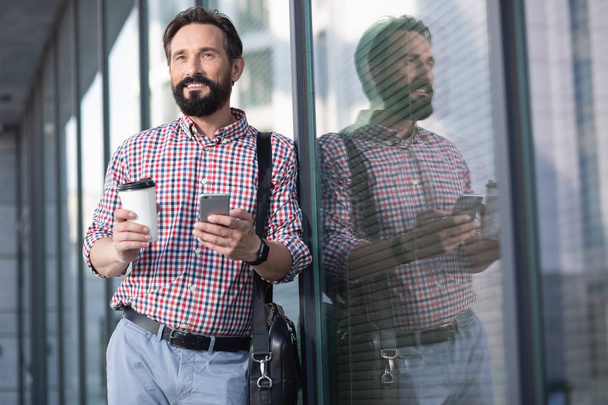 Busy days. Waist up of a pleasant bearded man drinking coffee while standing near office building - Fotoğraf, Görsel