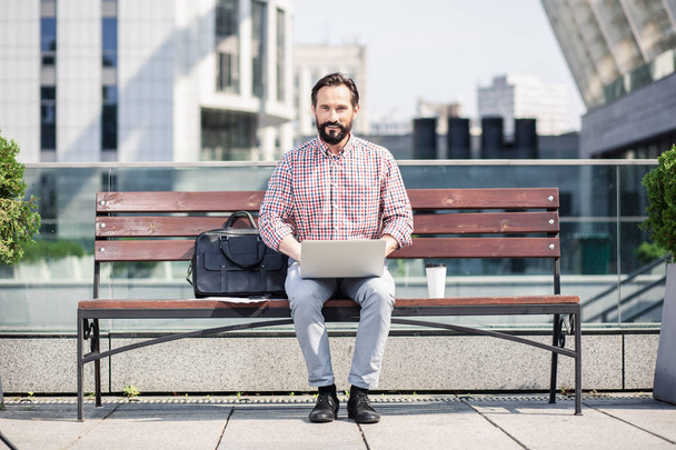 Ao ar livre. Agradável sorridente homem usando seu laptop enquanto sentado no banco
 - Foto, Imagem