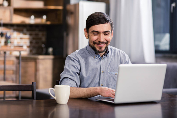 Online work. Cheerful handsome man using his laptop while enjoying distance work - 写真・画像