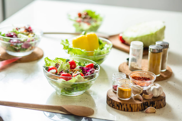 Close up of glass bowls with fresh vegetable salads and spices in glass bottles standing next to them on the pieces of wood - Foto, afbeelding