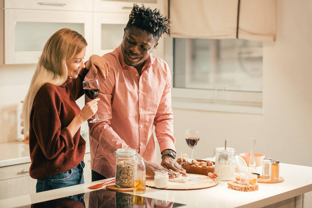 Стійка розслабленої молодої жінки, насолоджуючись червоним вином на кухні і поклавши одну руку на плече свого хлопця, спостерігаючи, як він замінює тісто
 - Фото, зображення