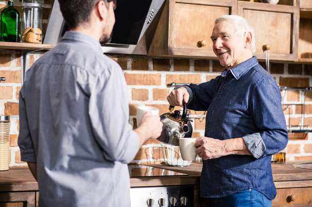 Warme sfeer. Vrolijke bejaarde man met een waterkoker terwijl hij thee drinkt met zijn zoon in de keuken - Foto, afbeelding