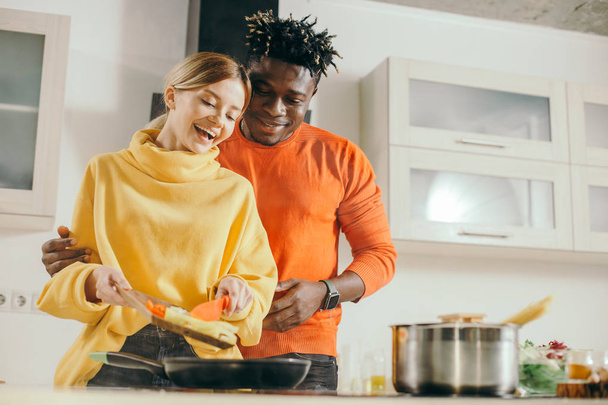 Excited young lady putting vegetables on the frying pan and smiling while cheerful boyfriend standing behind her back and watching the cooking process - 写真・画像