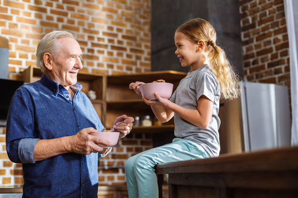 Try it. Joyful little girl enjoying her breakfast while talking with her grandfather - Photo, Image