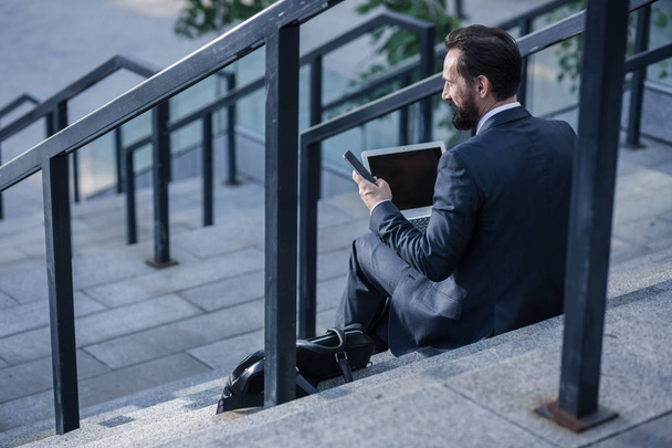 Get in touch. Pleasant professional businessman sitting on the stairs while using his smartphone - 写真・画像