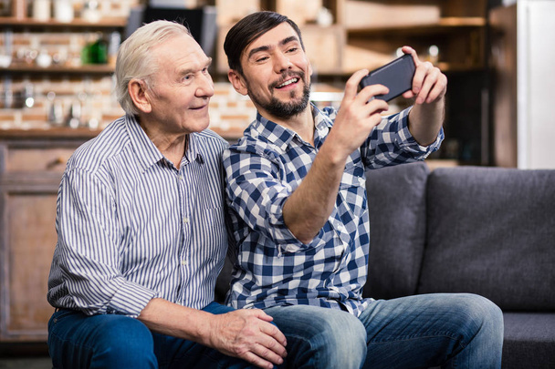 Just smile. Cheerful aged man sitting on the sofa with his son who is making selfies - Zdjęcie, obraz