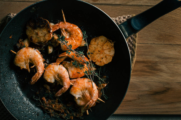 Big frying pan placed on the towel on wooden table. Fried tiger prawns with garlic and thyme on the pan - Фото, зображення