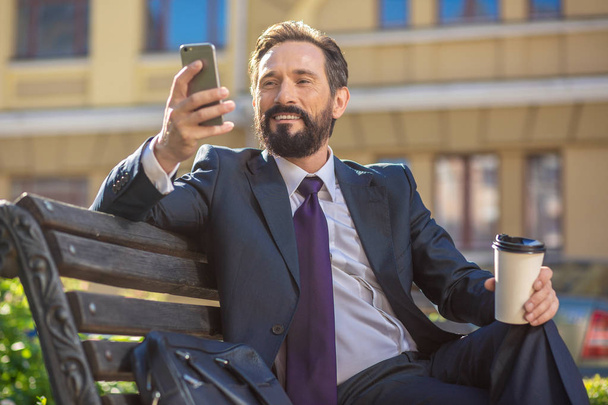 In a upbeat mood. Smiling bearded businessman sitting on the bench while resting with a coffee cup - Foto, Imagem