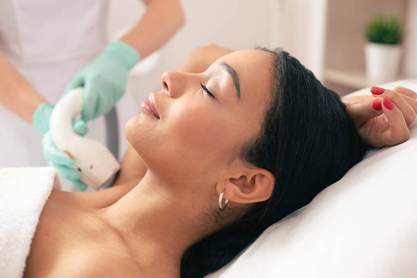 Young beautiful woman lying with her eyes closed and smiling while undergoing laser hair removal on her armpit - Foto, Bild