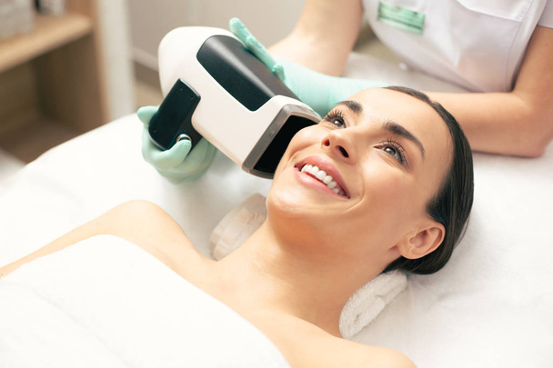 Positive young lady looking happy while skin analysis camera being placed near her face - Foto, Imagem
