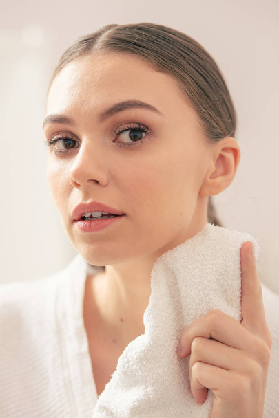 junge Frau mit natürlichem Make-up, die ihr Spiegelbild im Spiegel betrachtet, während sie ein Handtuch hält - Foto, Bild