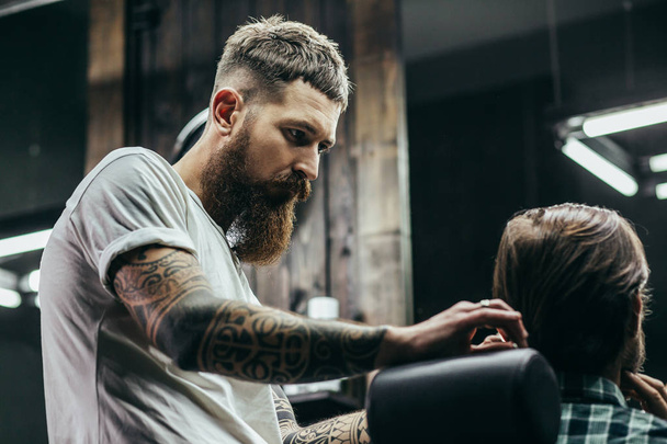 Внимательный концентрированный бородатый парикмахер, стоящий рядом с мужчиной и расчесывающий волосы
 - Фото, изображение