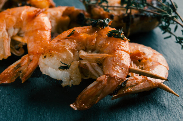 Two fried shrimp skewers with thyme sprigs lying on the black slate - Photo, Image