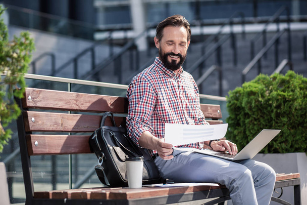 Freelance worker. Cheerful bearded man using his laptop while sitting on the bench - 写真・画像