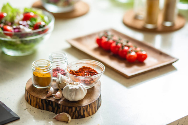 Selective focus of the spices in glass bottles standing on the piece of wood with garlic. Plate of cherry tomatoes and bowl of salad on the background - Photo, image