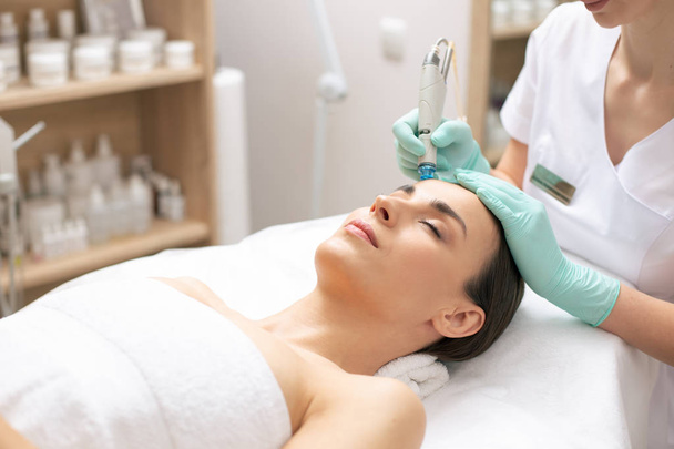 Calm young woman having her eyes closed while professional cosmetologist conducting dermabrasion procedure on her face - Photo, image