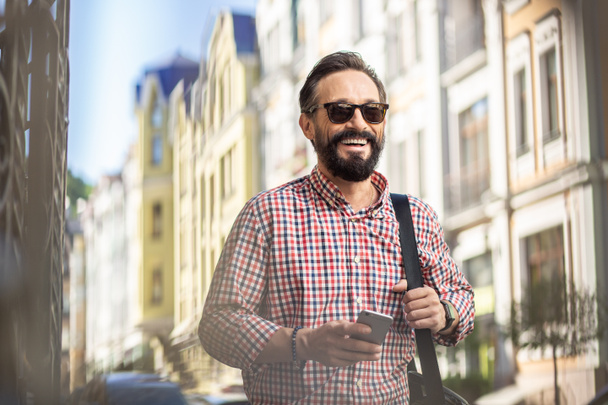 Użytkownik mobilny. Wesoły przystojny człowiek chodzenie wzdłuż The ulica podczas uśmiechnięty - Zdjęcie, obraz