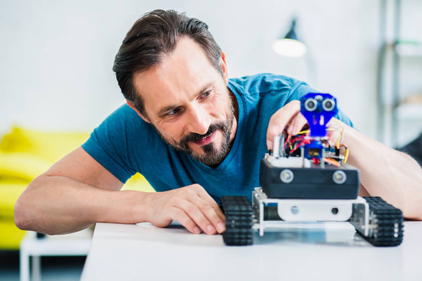 Overjoyed bearded man sitting at the table while constructing robot at home - 写真・画像