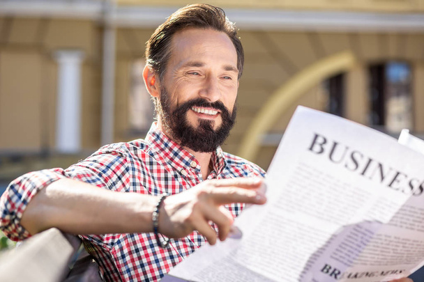 Pleasant morning. Cheerful adult man smiling while reading a daily newspaper - 写真・画像