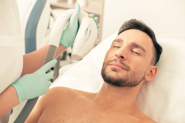 Dark haired bearded man on the white pillow closing his eyes during the photo rejuvenation procedure - Fotoğraf, Görsel