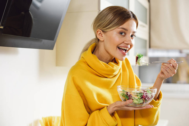 Positive young pretty lady in yellow sweater smiling and looking happy while eating delicious salad from the glass bowl - Фото, зображення