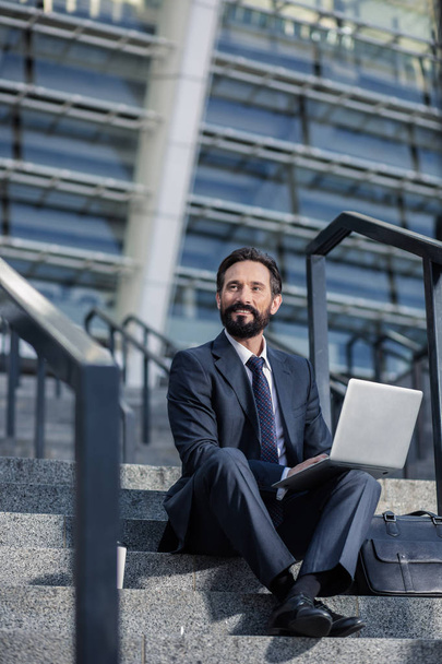 Professional mindset. Pleasant bearded businessman using his laptop on the staircases - Foto, imagen