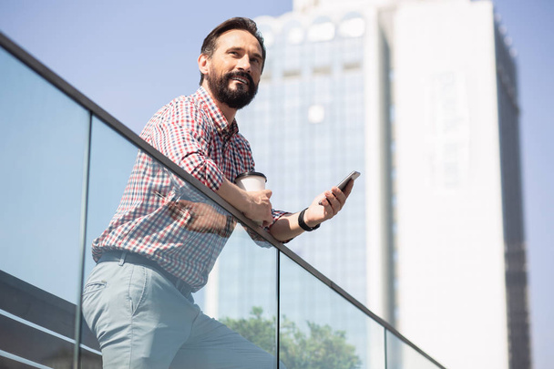 Confident look. Joyful bearded man using his phone while enjoying urban view - Foto, Bild