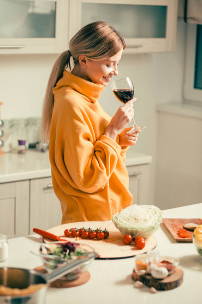 Cintura de calma jovem mulher em pé na cozinha ao lado da mesa com legumes sobre ele e sorrindo alegremente enquanto segurando um copo de vinho
 - Foto, Imagem