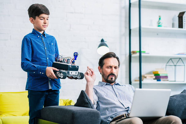 Ingenious schoolboy holding a robot while his busy father working online - Φωτογραφία, εικόνα