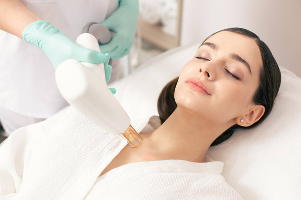 Dark haired lady lying with closed eyes while a professional medical worker holding a modern tool and removing scars with a laser - Foto, Imagem