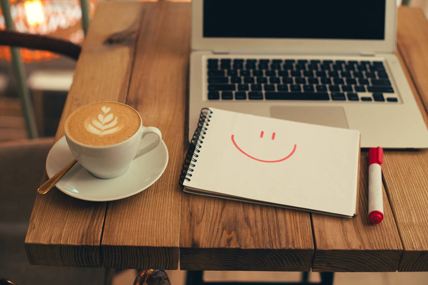 Notebook met een getekend glimlachend gezicht geplaatst op de houten tafel tussen een laptop en een kopje koffie - Foto, afbeelding