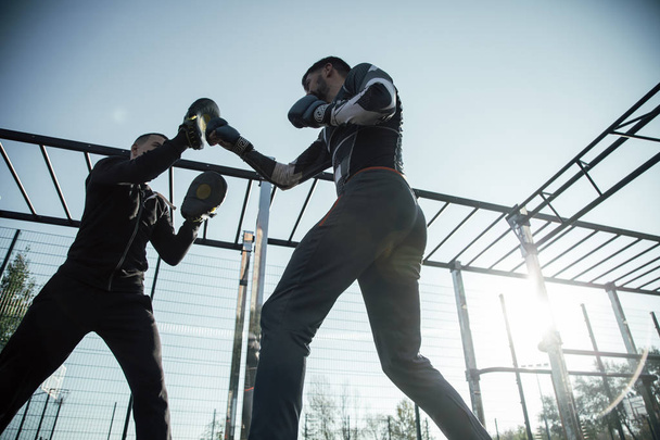 Laconic photo of young MMA boxer standing at the sports ground with his trainer and practicing punches on focus mitts - Fotó, kép