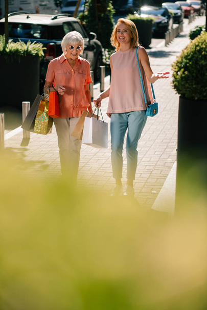 Gladsome volwassen vrouw en haar moeder lopen bij zonnig weer. Kleurrijke papieren zakken in hun handen - Foto, afbeelding