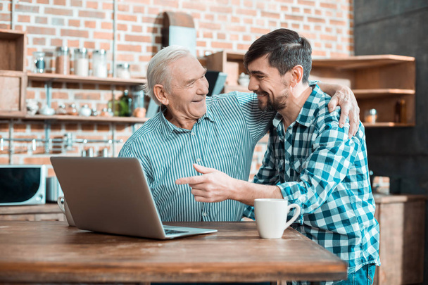 Warm relationships. Cheerful nice elderly man smiling and hugging his son while learning how to use a laptop - Foto, immagini