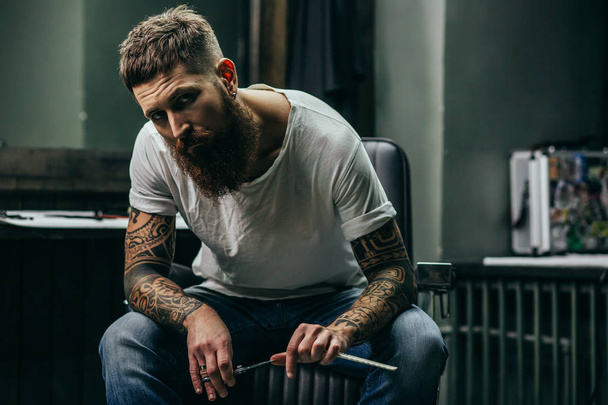 Calm bearded tattooed man sitting in the armchair and frowning while holding straight razor - Foto, Imagem