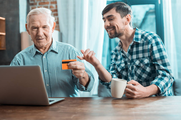 New technologies. Delighted positive young man smiling and pointing at the laptop screen while helping his grandfather to do an online payment - Фото, изображение
