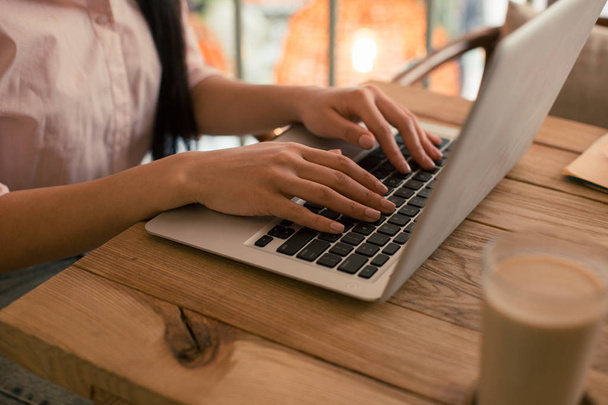 Close-up foto van een houten tafel met een kopje koffie in de buurt van de laptop en vrouw handen op het toetsenbord - Foto, afbeelding