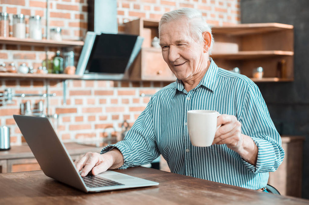 Modern technology. Delighted positive aged man smiling and holding a cup of tea while using a laptop - 写真・画像